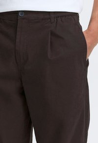 Mørkebrune bukser med flad front, der har en knap og elastisk talje, sidelommer og en glat bomuldsstruktur.