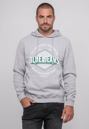 Jeune homme aux cheveux courts portant un sweat à capuche gris clair avec l'inscription "Blue & Jeans" sur le devant, debout avec une main dans la poche.