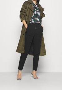 Manteau trench olive, blouse noire à fleurs avec pois blancs, pantalon noir et talons strappy bleus. Tissu lisse, coupe ajustée.