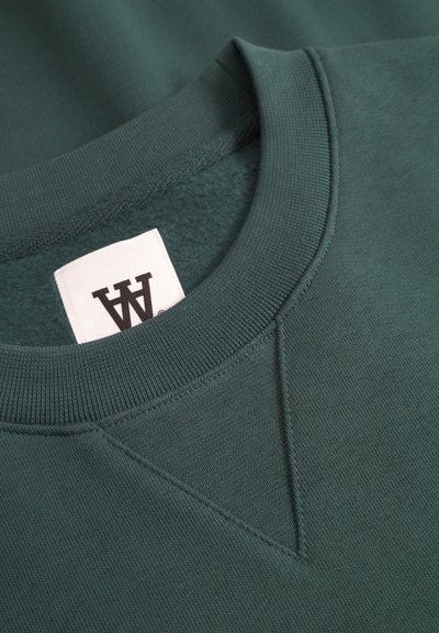 Sweatshirt vert foncé avec un col rond à bord côtelé, en tissu texturé, et une petite étiquette blanche avec impression noire.