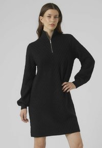 Abito nero lavorato a maglia con collo a lupetto con zip, maniche a sbuffo lunghe e motivo a zigzag testurizzato. L'orlo arriva a metà coscia.