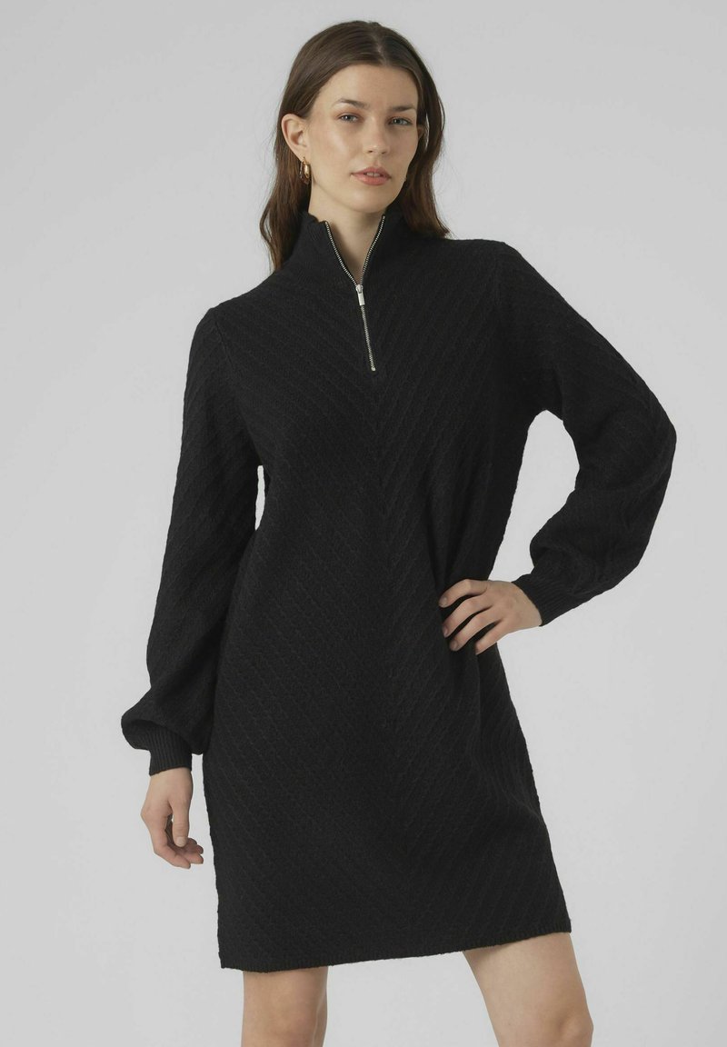 Abito nero lavorato a maglia con collo a lupetto con zip, maniche a sbuffo lunghe e motivo a zigzag testurizzato. L'orlo arriva a metà coscia.