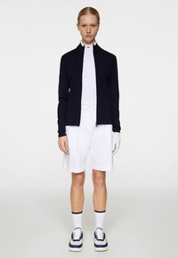 Tumši zila jaka ar rāvējslēdzēju, balta īsroku krekls, balti šorti un sporta apavi ar ziliem akcentiem; roka ir pārklāta ar cimdu.