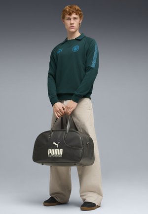 Junger Mann in dunkelgrünem Sweatshirt und beigen Hosen, der eine schwarze Puma-Reisetasche hält und vor einem grauen Farbverlauf-Hintergrund steht.