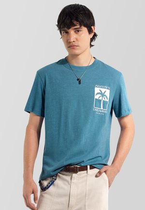 Junger Mann trägt ein türkisfarbenes T-Shirt mit weißem Palmenmotiv und dem Schriftzug "Always Dreaming", beige Hose, braunen Gürtel und Anhänger-Halskette.