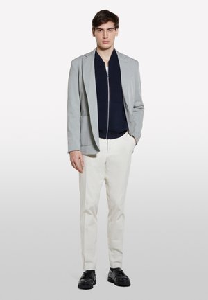 Blazer gris clair au design structuré, sweat-shirt zippé marine en dessous, pantalon crème et chaussures noires, se tenant devant un fond blanc.