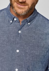 Behaarde man met een blauw overhemd met knopen en witte knopen, zichtbaar in de kraag en het bovenste deel van de borst.