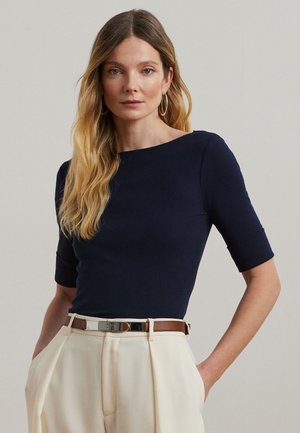 Ženska z dolgimi blond lasmi, ki nosi modrikasto mornarsko oprijet zgornji del s komolčnimi rokavi in krem hlače z visokim pasom ter rjavim pasom.