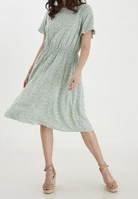 Robe à imprimé floral vert avec des manches courtes, taille froncée et jupe fluide. Portée avec des sandales compensées beiges. Texture lisse.