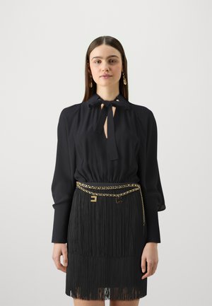 Blusa nera con colletto a cravatta, dotata di maniche lunghe. Abbinata a una gonna nera con frange, accentuata da una cintura con catena dorata.