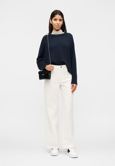 Γυναίκα που φοράει μπλε σκούρο πουλόβερ πάνω από ριγέ ζιβάγκο, άσπρο παντελόνι με φαρδιά πόδια, άσπρα αθλητικά παπούτσια, κρατώντας μαύρη τσάντα χιαστί.