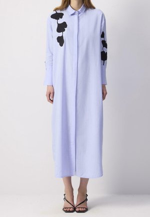 Femme portant une longue robe-chemise bleu clair avec des motifs de feuilles noires sur l'épaule et la manche, associée à des talons noirs à brides.