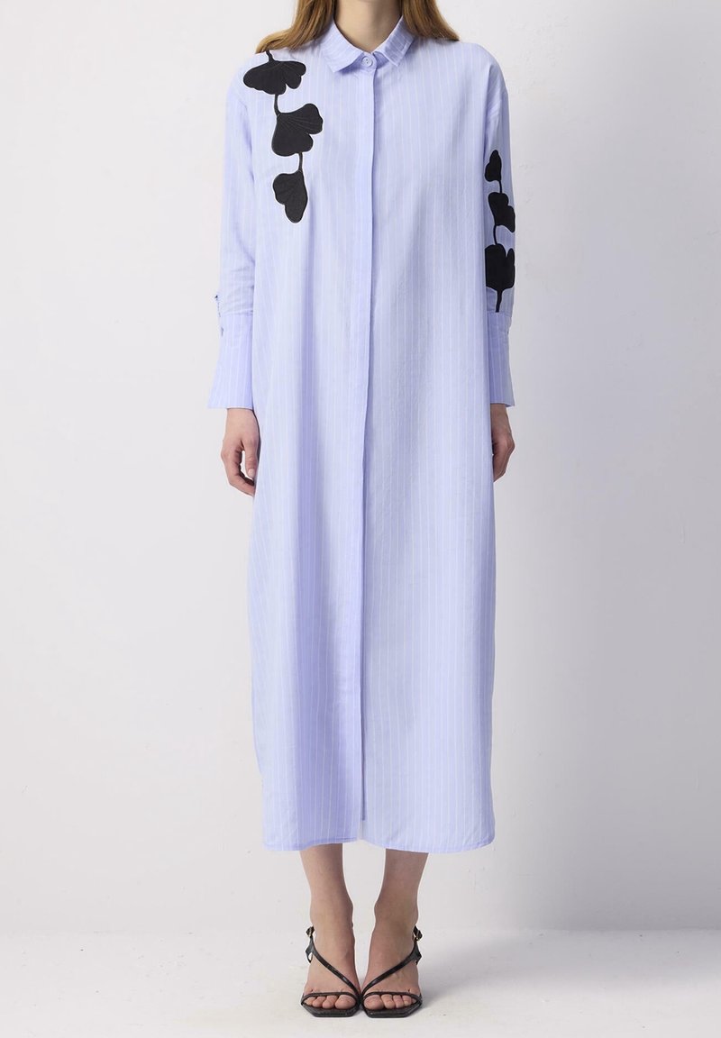 Femme portant une longue robe-chemise bleu clair avec des motifs de feuilles noires sur l'épaule et la manche, associée à des talons noirs à brides.