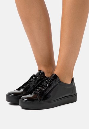 Nogi w błyszczących czarnych niskich trampkach ze sznurowadłami, gładkim wykończeniem i grubymi czarnymi podeszwami na jednolitym jasnym tle.