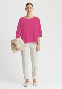 Fuchsfarbenes Übergrößen-Oberteil mit Rundhalsausschnitt, kombiniert mit hellbeigen maßgeschneiderten Hosen mit umgeschlagenen Säumen und silbernen Slippern.