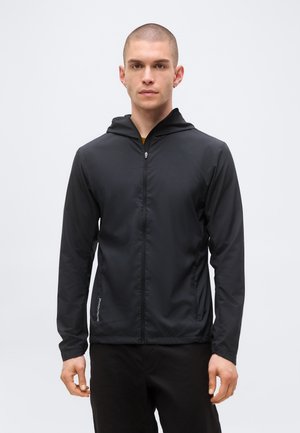 Mladi muškarac s kratkom kosom, obučen u crnu jaknu s kapuljačom i patentnim zatvaračem i crne hlače, stoji ispred jednobojne svijetle pozadine.