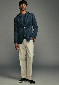 Blazer en daim marine, à un bouton, avec revers crantés, associé à un haut bleu et un pantalon beige clair. Chaussures noires à enfiler, texture lisse.
