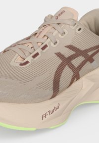 Κοντινό πλάνο μπεζ αθλητικού παπουτσιού ASICS με ανοιχτό ροζ λεπτομέρειες, καφέ λογότυπο, μπεζ κορδόνια, ετικέτα FF Turbo2 στη σόλα και λάιμ πράσινη άκρη εξωτερικής σόλας.
