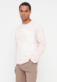 Camicia a maniche lunghe di un delicato rosa chiaro con un ampio motivo floreale bianco, realizzata in un tessuto morbido; abbinata a pantaloni beige a coste.