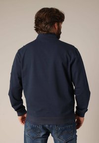 Marineblauwe sweatshirt met hoge kraag, ribgebreide manchetten en zoom, en gestikte schouderdetails, gedragen met blauwe jeans.