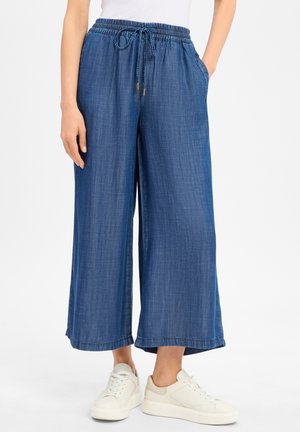 Kvinde iført løse blå denim bukser med vide ben, elastisk linning og hvide sneakers.