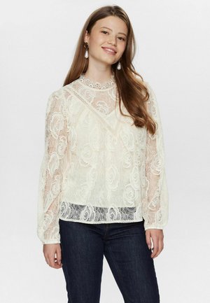 Cremefarbige Spitzenbluse mit floralem Muster, hohem Kragen und langen Puffärmeln; kombiniert mit dunkelblauen Jeans.