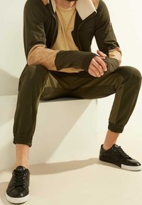 Chándal de color verde oliva con detalles en ante color marrón claro en los hombros, mangas largas y puños elásticos. Zapatillas negras con suelas blancas completan el look.