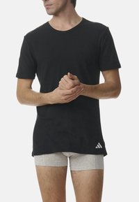 T-shirt nera in cotone con maniche corte, scollatura rotonda e logo Adidas grigio in basso. Sotto, si vedono pantaloni boxer grigi chiari.