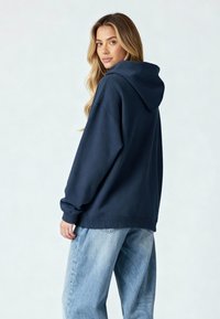 Felpa oversized blu navy con polsini e orlo a coste, dotata di cappuccio e texture morbida, abbinata a jeans baggy in denim chiaro.