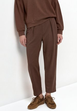Persona in piedi che indossa pantaloni marroni a taglio largo, maglia a maniche lunghe marrone coordinata e sneakers marroni in camoscio con lacci su sfondo bianco.