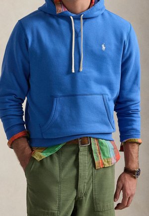 Homme portant un sweat à capuche bleu par-dessus une chemise à carreaux multicolores, un pantalon vert avec des poches et une montre-bracelet, mains dans les poches.