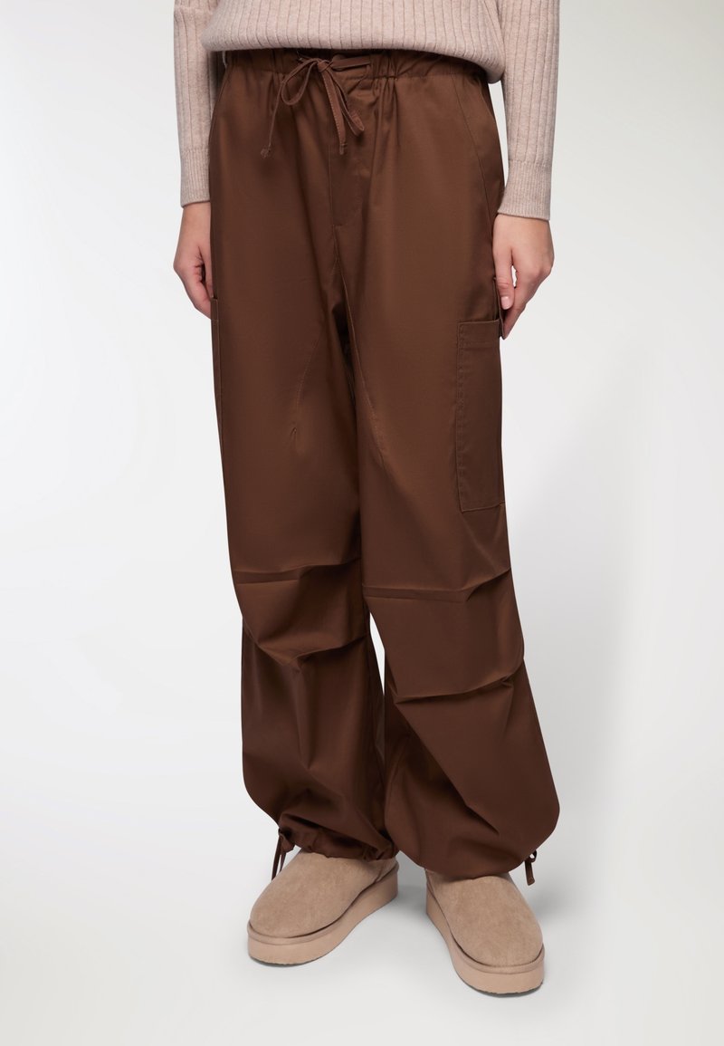 Pantaloni marroni, larghi e comodi, realizzati in un tessuto liscio, con una cintura elastica e un cordoncino, e con tasche laterali.