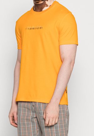 Pomarańczowa bawełniana koszulka z okrągłym dekoltem i wytłaczanym logo „Calvin Klein” na przodzie. Krótkie rękawy i luźny krój.