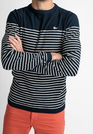 Uomo con le braccia incrociate che indossa un maglione a maniche lunghe a righe blu navy e bianche e pantaloni rossi su sfondo bianco uniforme.
