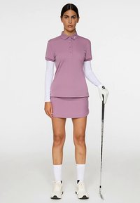 Golfoutfit met een lichtpaarse polo met een kraag en drie knopen, gecombineerd met een bijpassende rok. Witte lange mouwen en handschoenen.