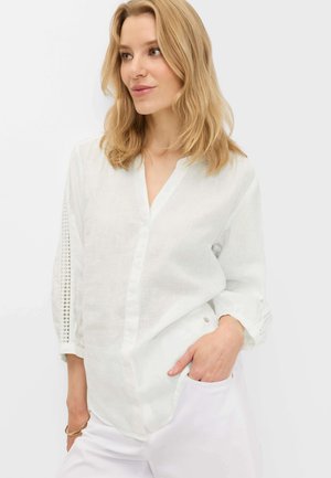 Femme blonde portant un chemisier blanc texturé avec des détails en dentelle aux manches, main dans la poche, regardant légèrement sur le côté.