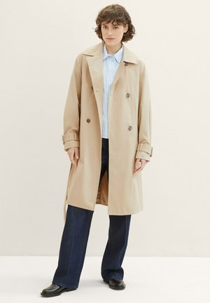 Beige dobbeltradet trenchcoat med store brune knapper, skulderremme og en løs pasform, båret over en lyseblå stribet skjorte og mørke jeans.