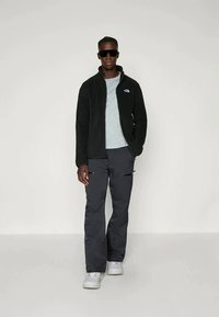 Melna vilnas jaka ar priekšējo rāvējslēdzēju, gaiši zils T-krekls, tumši pelēkas cargo bikses ar rāvējslēdzēja kabatām un baltas kurpes. Valkā saulessargus.