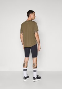 Olivenfarbendes Kurzarmhemd mit Rundhalsausschnitt, kombiniert mit schwarzen Shorts und weißen Socken mit schwarzen Streifen, getragen mit schwarzen Sneakern.