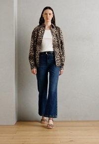 Løveprint knap-op skjorte, hvid t-shirt, flagrende blå jeans og mønstrede sandaler, placeret i et minimalistisk rum med trægulv.