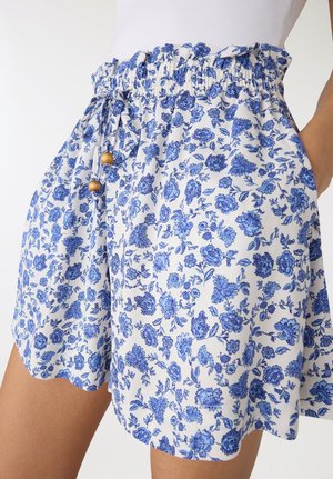 Pantaloncini bianchi con stampa floreale blu, vita elastica con coulisse e perline di legno, indossati con una mano in tasca.
