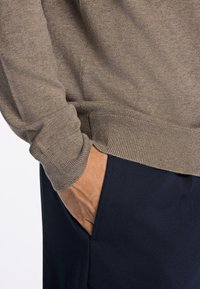 Brązowy sweter z dzianiny z prążkowanymi mankietami, zestawiony z granatowymi spodniami z boczną kieszenią. Zbliżenie na dłoń spoczywającą w kieszeni.