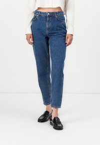 Jeans de denim azul de tiro alto con un ajuste relajado, que cuentan con una cremallera, pasadores para cinturón y piernas ligeramente ajustadas. Llevados con mocasines negros.