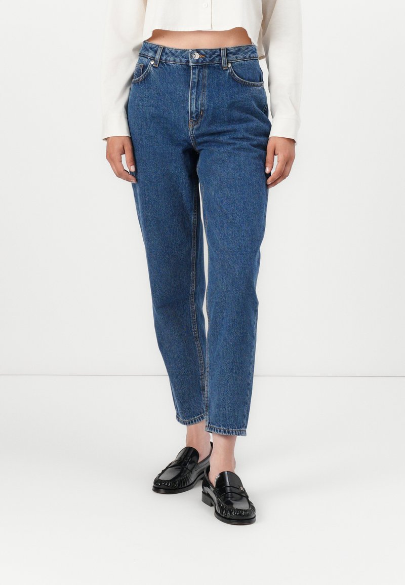 Jeans de denim azul de tiro alto con un ajuste relajado, que cuentan con una cremallera, pasadores para cinturón y piernas ligeramente ajustadas. Llevados con mocasines negros.