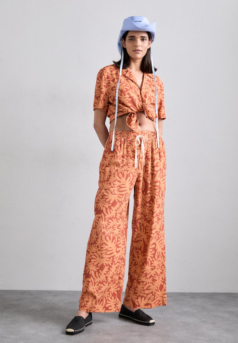 Orange patterned crop top tied at the waist, paired with wide-leg pants. Light blue bucket hat with strings and black espadrilles. Smooth texture.