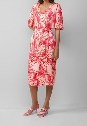 Kobieta ubrana w sukienkę kopertową o długości do połowy łydki, w różowo-czerwony kwiatowy wzór, z marszczonymi rękawkami i różowe sandały na obcasie z paskami, na jednolitym szarym tle.