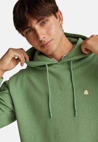 Felpa verde con cappuccio regolabile, con un piccolo logo giallo sul petto. Il tessuto sembra morbido e con una trama liscia.