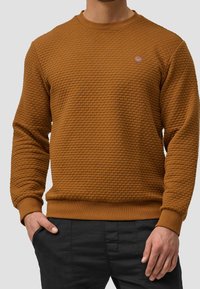 Brązowa bluza z teksturą, z dekoltem w kształcie łódki, posiadająca prążkowany kołnierz i mankiety oraz małe logo na piersi.