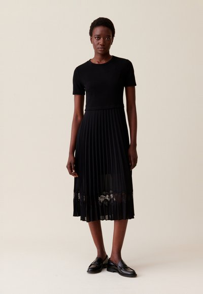 Black pleated dress with short sleeves, featuring a lace detail at the hem. Worn with black loafers, simple design, smooth texture.