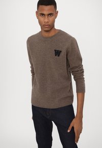 Brown knitted sweater with a crew neckline and cuffed sleeves; features a black "W" logo on the left chest; paired with dark jeans.